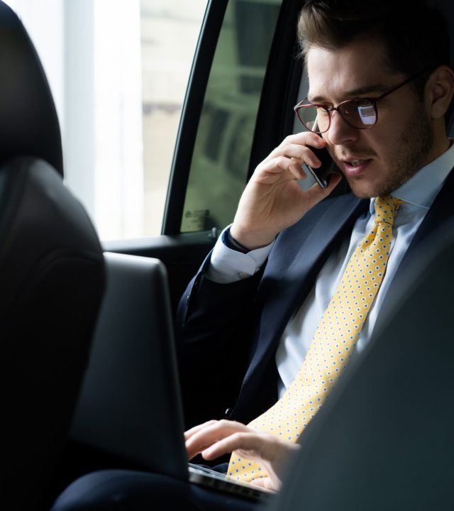 Young businessman sitting on back seat of the car, while his chauffeur is driving automobile